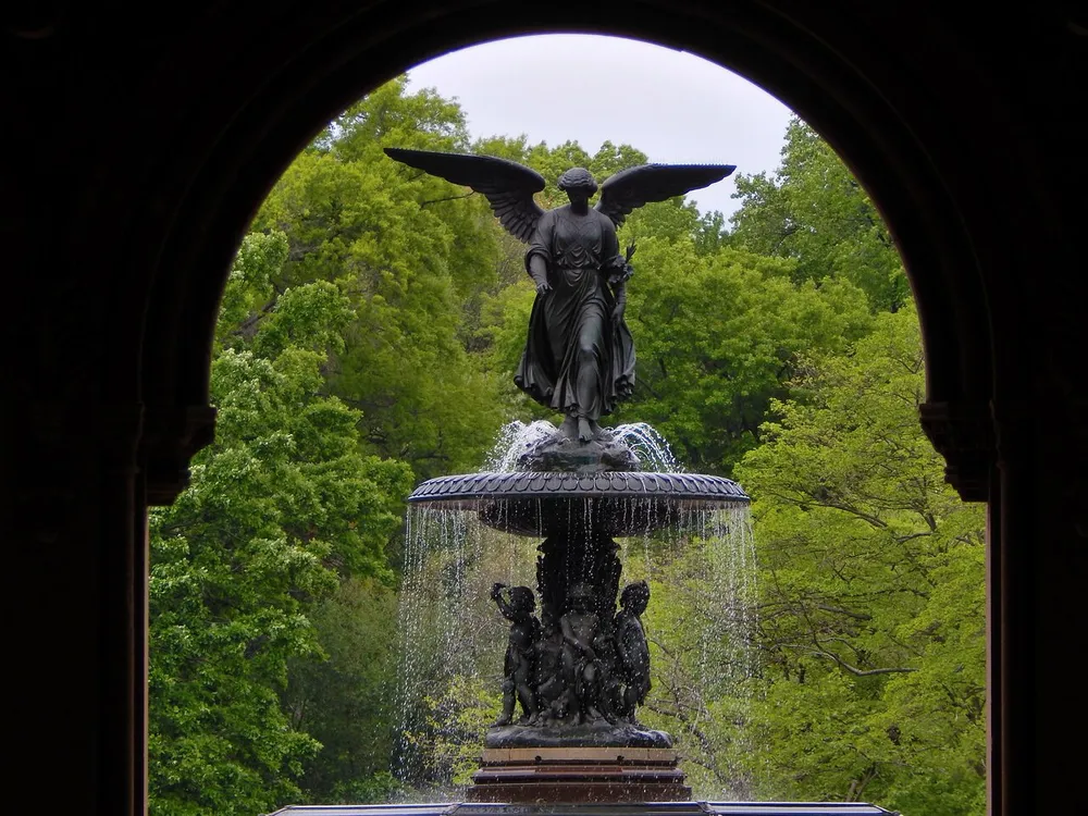 The image shows a view of an angelic figure atop a fountain framed by an ornate archway with lush green trees in the background