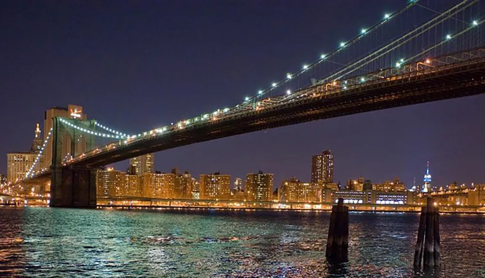 The image shows the illuminated Brooklyn Bridge at night with the East River in the foreground and the city skyline in the background