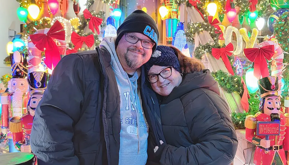Two people are smiling for a photo in front of a vibrant Christmas-themed decoration that includes lights nutcrackers and festive bows