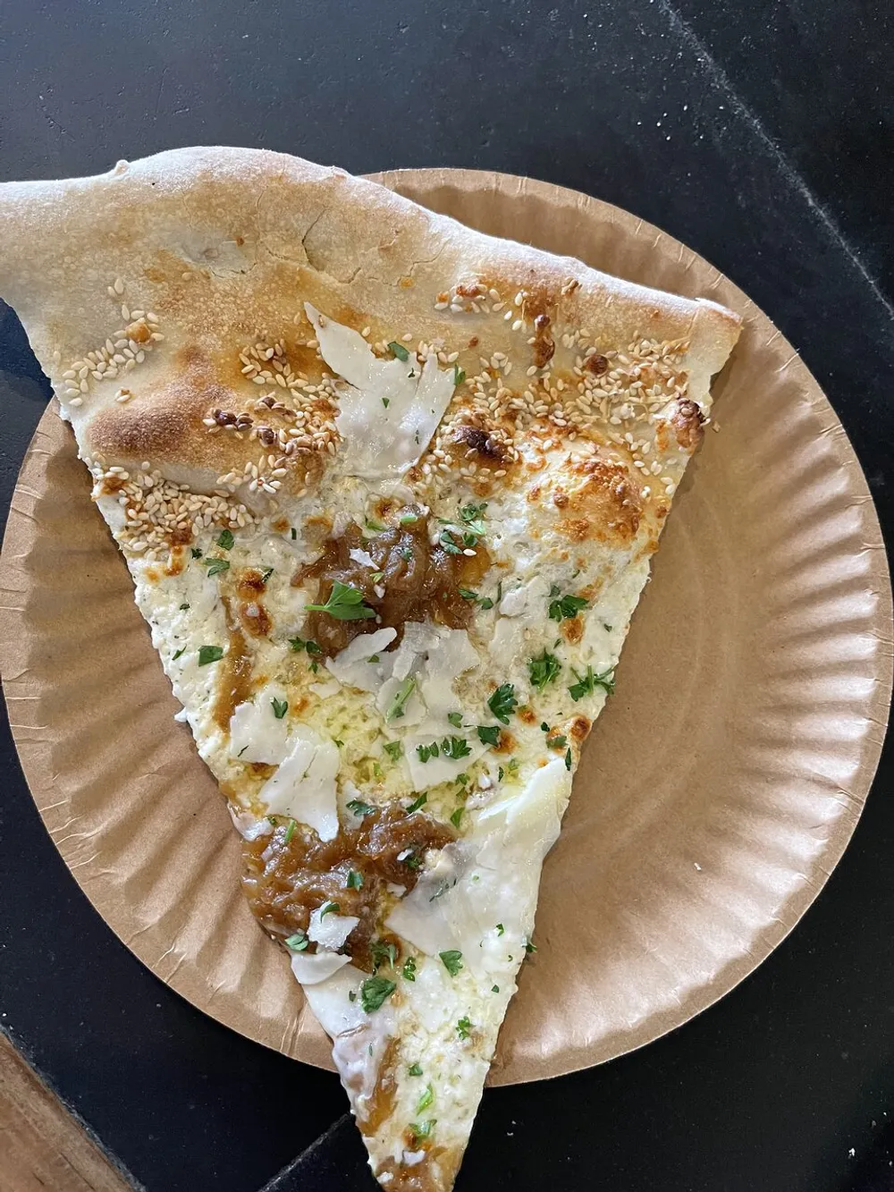 The image shows a slice of pizza with white cheese and caramelized onions sprinkled with sesame seeds and herbs on a brown paper plate