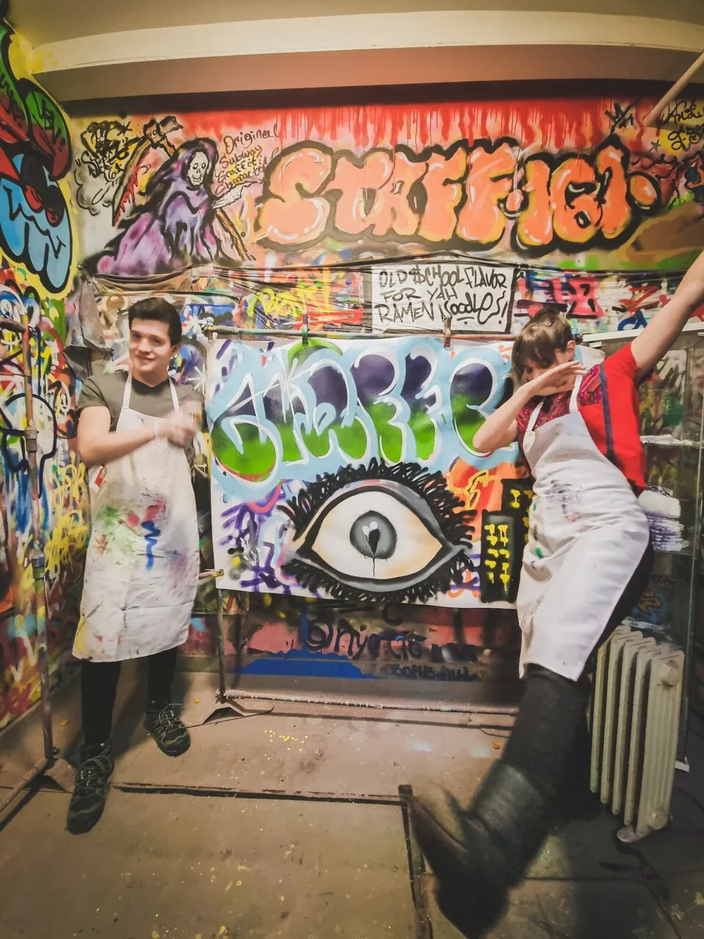 Two people in aprons are surrounded by vibrant graffiti art on walls with one person pointing at the camera and the other striking a playful pose