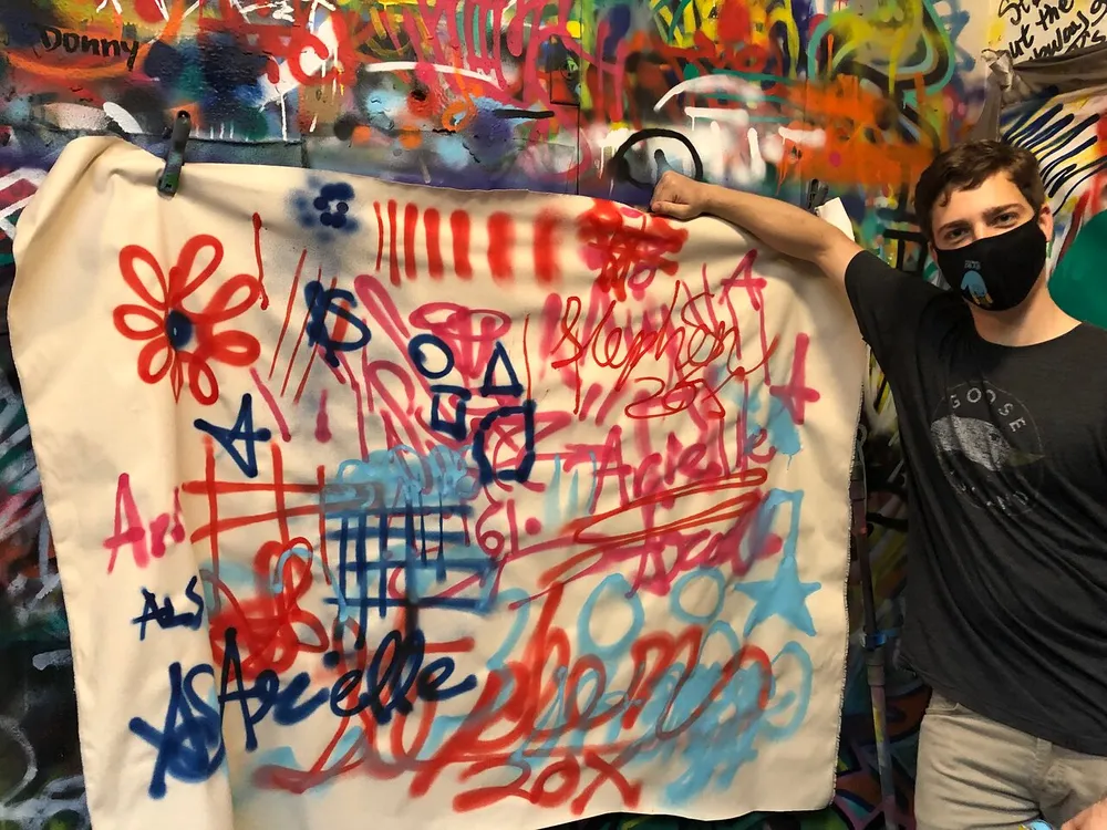 A person with a face mask is holding up a piece of fabric covered in colorful graffiti in front of a similarly tagged wall