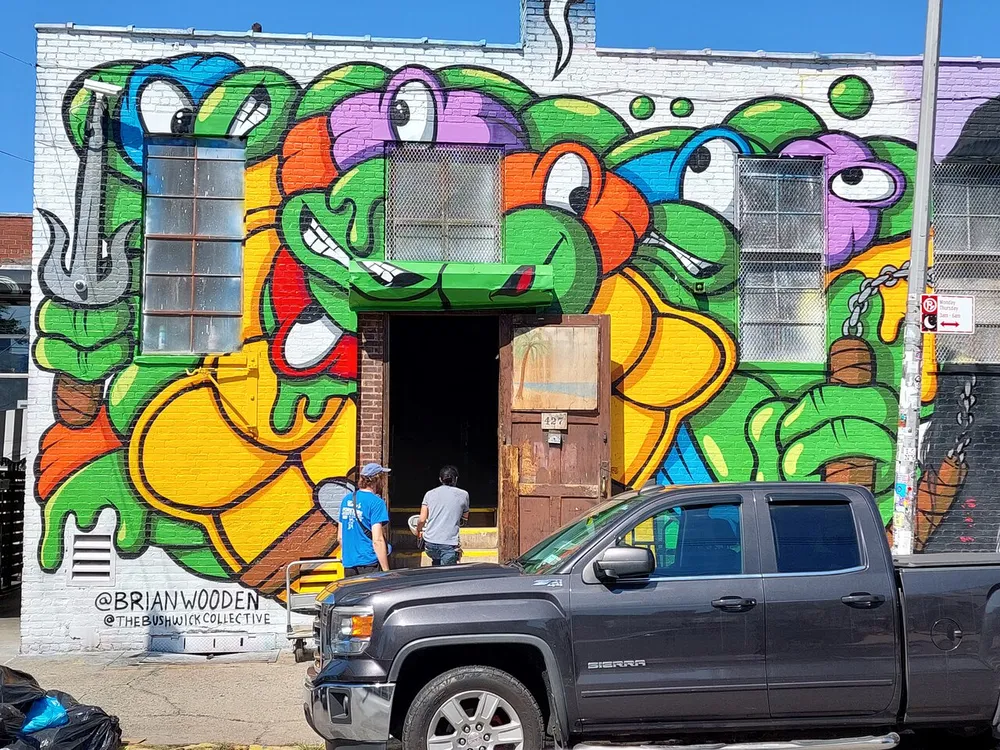 The image shows a vibrant street mural featuring colorful cartoonish snake characters on a buildings exterior with two people and a truck visible in front of the artwork