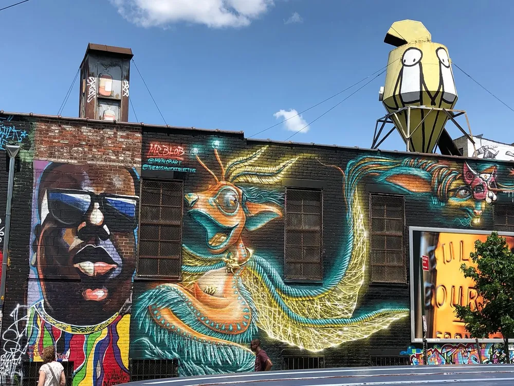 The image shows a vibrant street art mural featuring various colorful and stylized figures with a whimsical sculpture on a water tower above the building