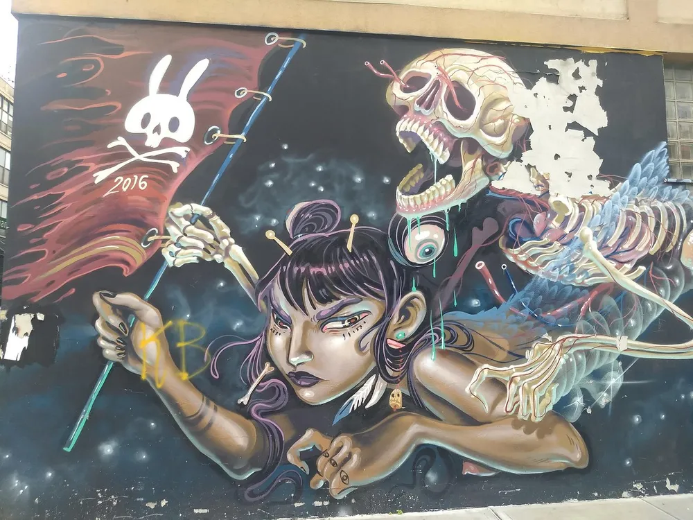 This is a vibrant street art mural depicting a woman with puppet strings controlling a skeleton and a skull marked with a bunny symbol blending elements of fantasy and surrealism