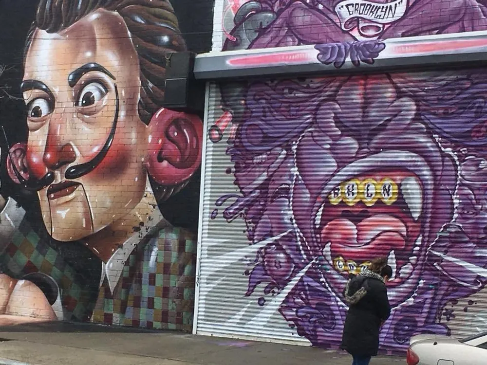 A person is walking past colorful street art murals on urban building shutters one of which resembles a clown-like face