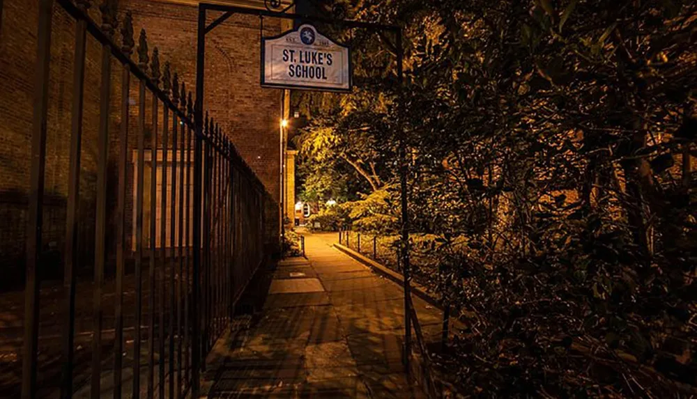 The image shows a warmly lit nighttime scene of an alleyway alongside St Lukes School bordered by an iron fence and overgrown greenery