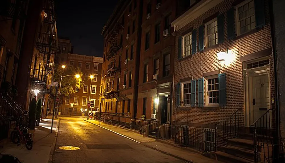 This image depicts a quiet dimly lit urban street at night showcasing the warm glow of street lamps on classic brick buildings with shuttered windows
