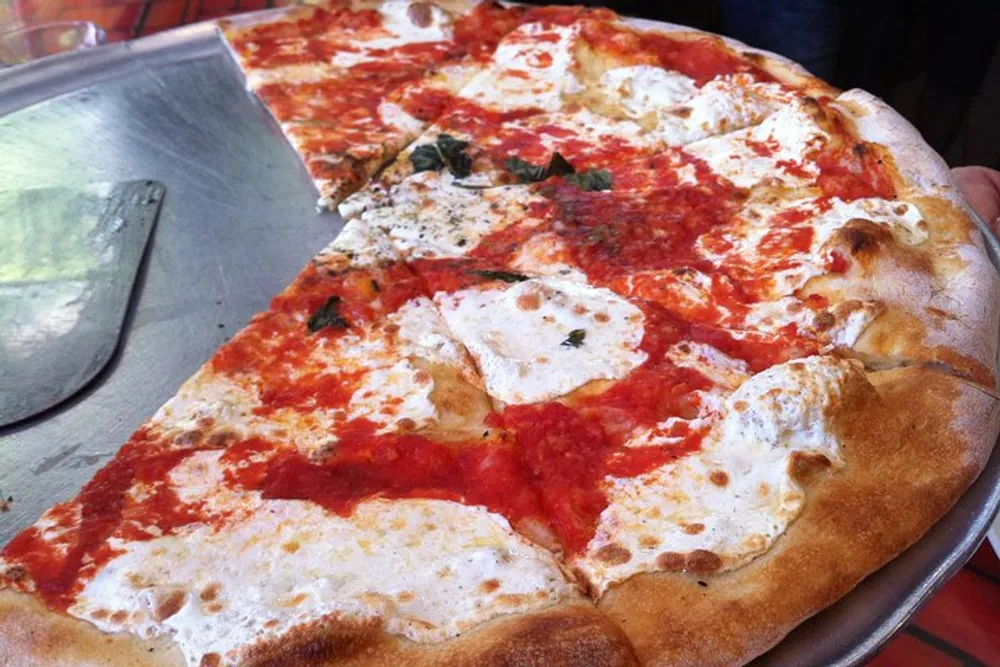 A large Margherita pizza with slices missing is displayed on a table with a pizza peel spatula on the side