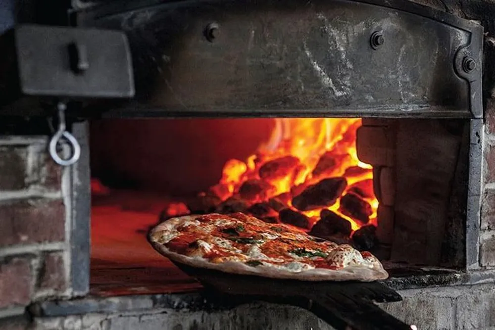 A pizza is being cooked in a wood-fired oven showcasing the flames in the background that provide the heat