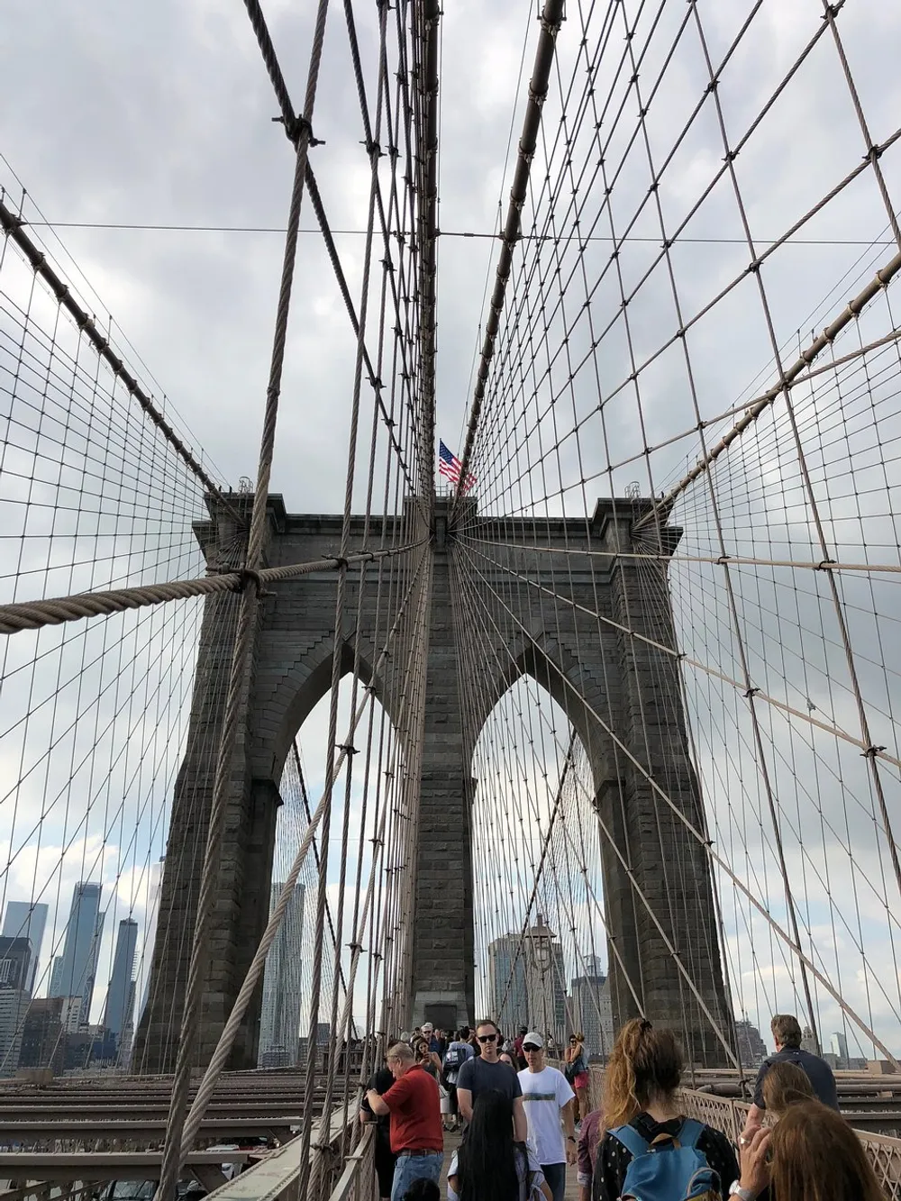 Visitors walk along the pedestrian pathway of the Brooklyn Bridge with its distinctive cable design framing the American flag and the New York City skyline in the distance