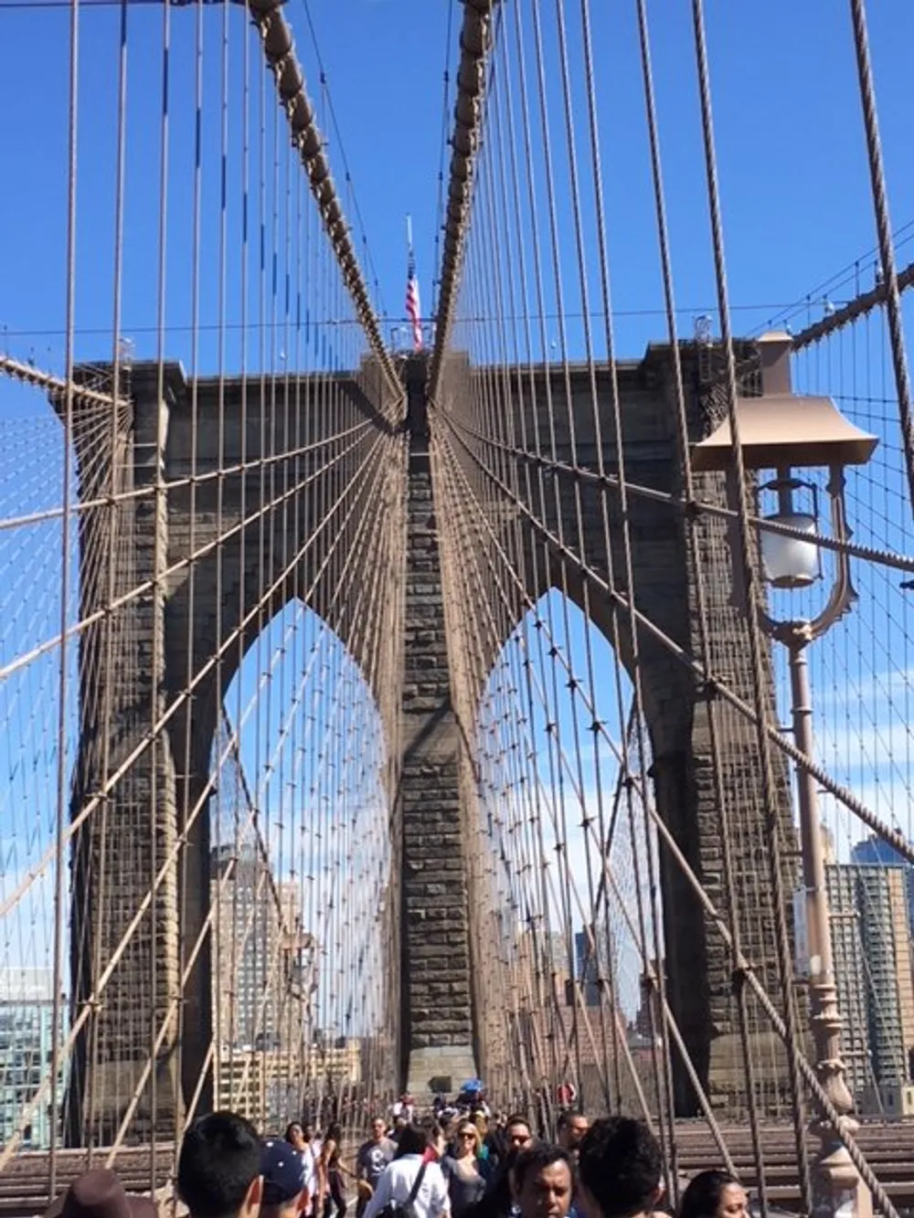 The image shows pedestrians walking along the walkway of the Brooklyn Bridge framed by its iconic suspension cables and stone towers against a clear blue sky
