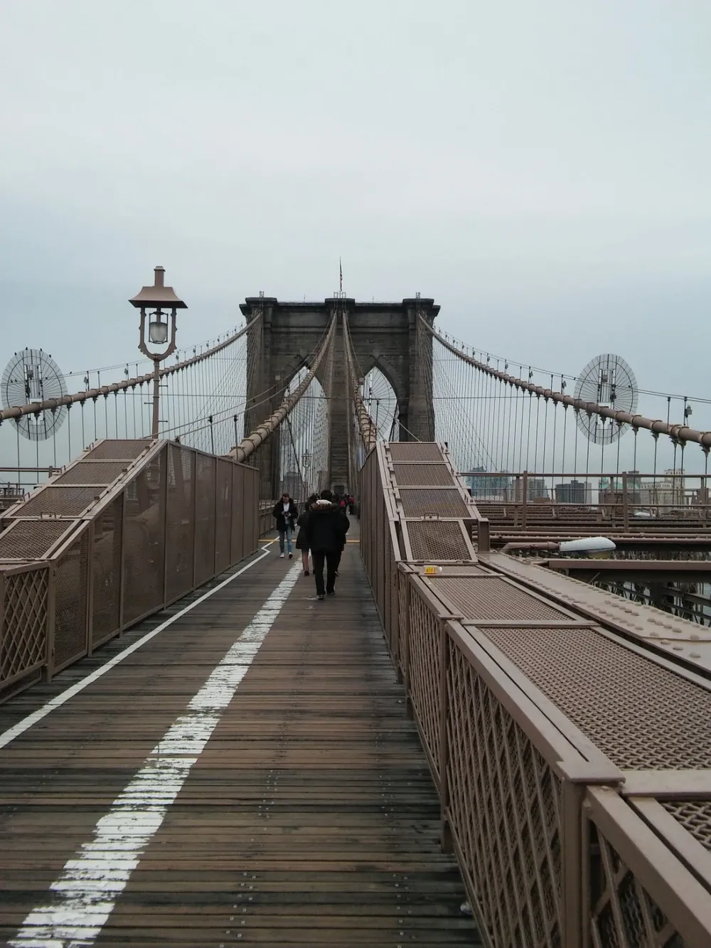 The image shows pedestrians walking on the wooden pedestrian pathway of a large suspension bridge possibly on an overcast day
