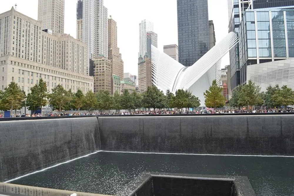 This image features one of the 911 Memorial pools with cascading waterfalls in the foreground the distinctive white wings of the Oculus structure in the background and surrounding skyscrapers all under a cloudy sky