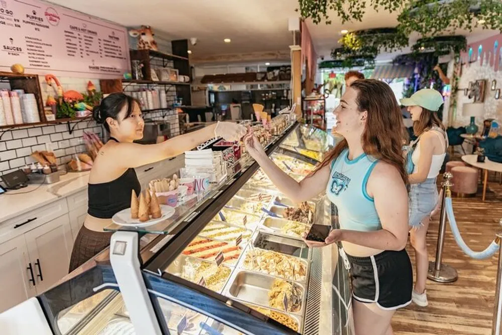 A customer at an ice cream shop is receiving a cone from the server while other patrons browse the selection