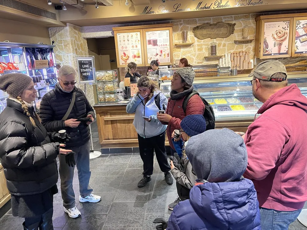 A group of people dressed warmly are waiting in line at a gelateria with some engaged in conversation and others looking at their phones
