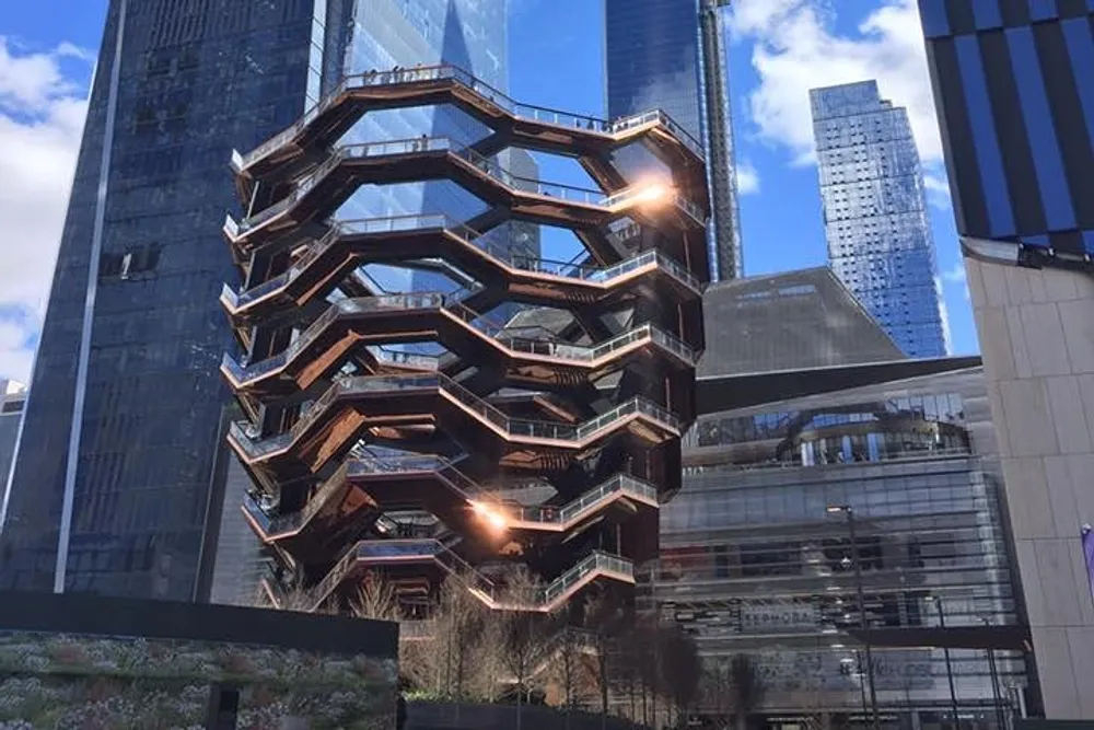 The image shows the Vessel a honeycomb-like structure of interconnected staircases located in Hudson Yards New York City surrounded by high-rise buildings under a clear blue sky