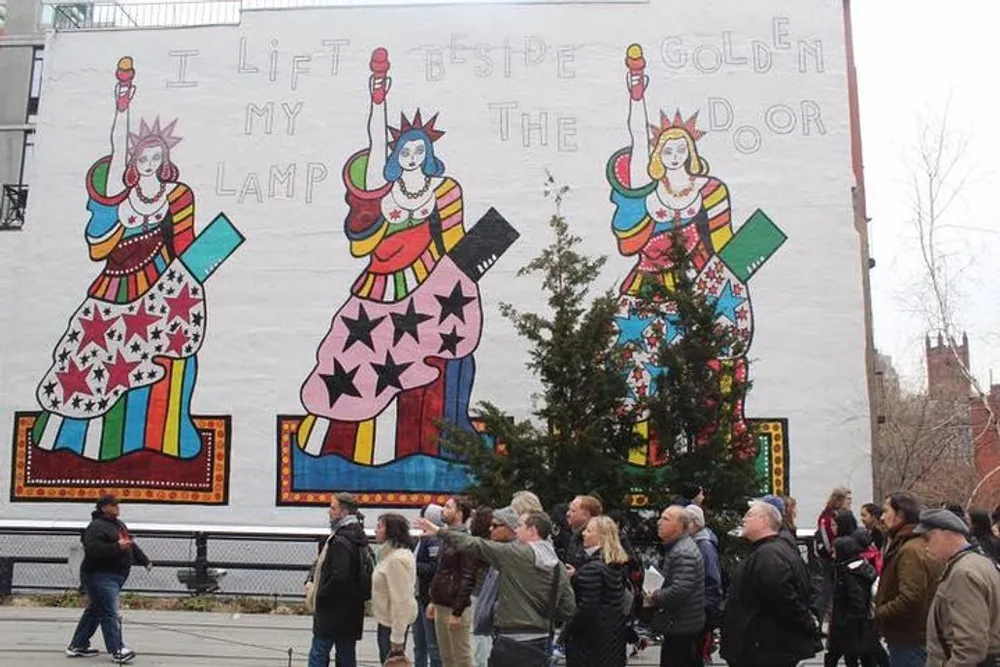 A group of people is observing a large mural depicting two stylized figures reminiscent of the Statue of Liberty with a phrase partially visible that appears to be inspired by the poem The New Colossus against the background of a large white wall
