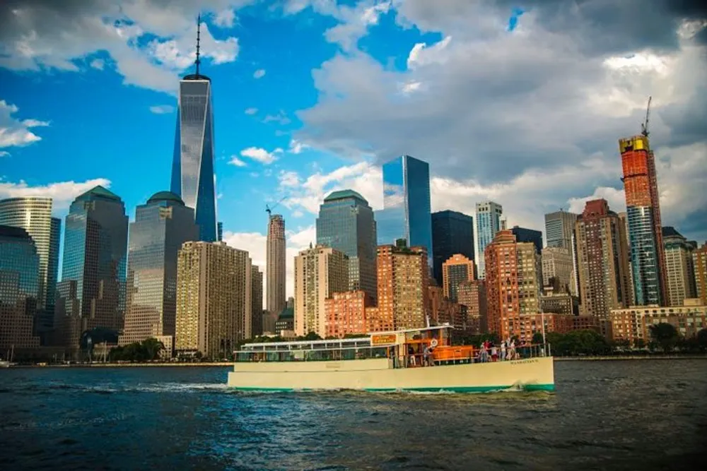 A ferry boat filled with passengers cruises along the water with a backdrop of the Manhattan skyline featuring the One World Trade Center under a partly cloudy sky