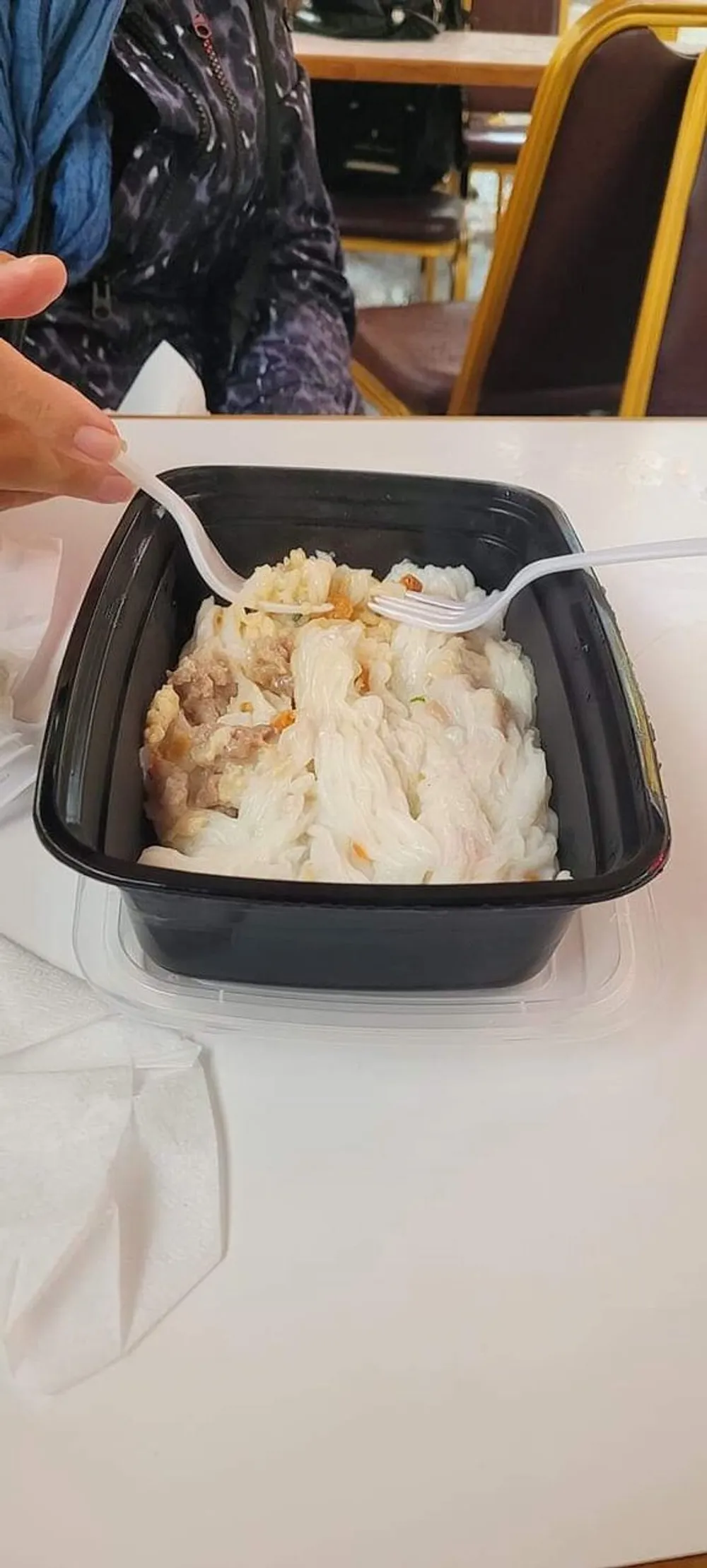 A person is about to eat a meal of rice and what appears to be chicken from a black plastic takeout container using a white plastic fork