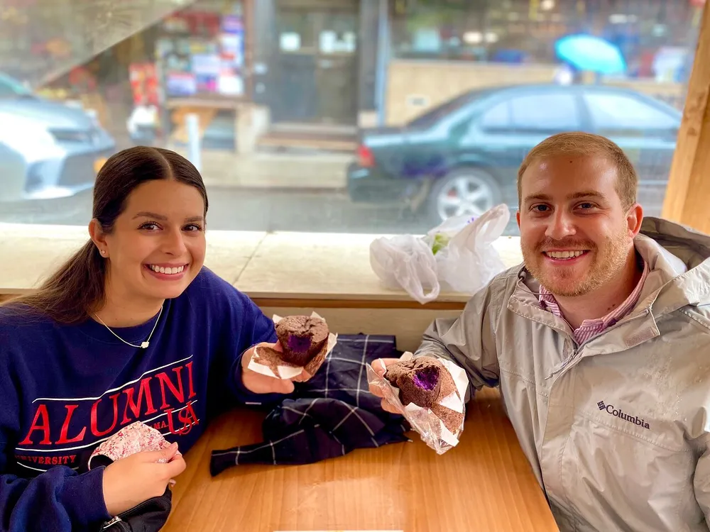 Two people are smiling at the camera while holding muffins seated inside a cafe with a street view through a large window behind them