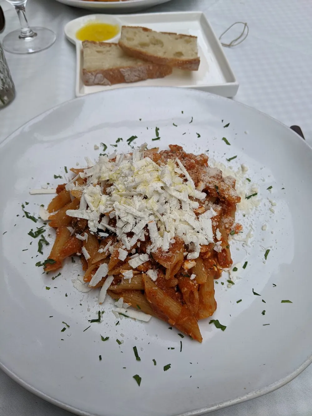The image shows a plate of pasta topped with grated cheese accompanied by slices of bread and olive oil served on a white plate at a dining table