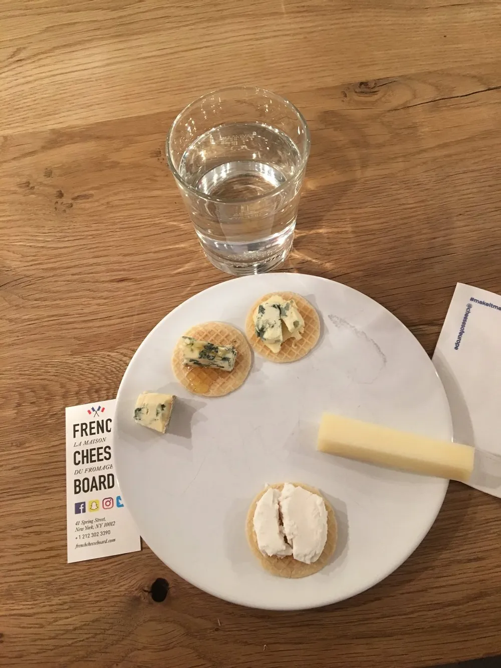 The image shows a water glass and a plate with different types of cheese on crackers alongside a cheese knife and a card from the French Cheese Board