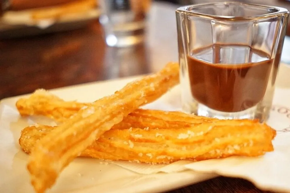 A plate of fresh churros is accompanied by a glass of chocolate sauce for dipping