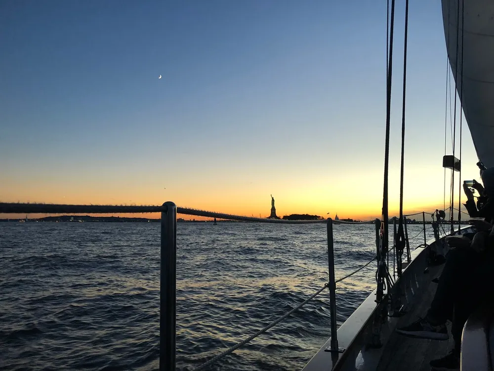 The image captures a serene sunset view from the deck of a sailboat with the Statue of Liberty silhouetted in the distance and a crescent moon in the twilight sky
