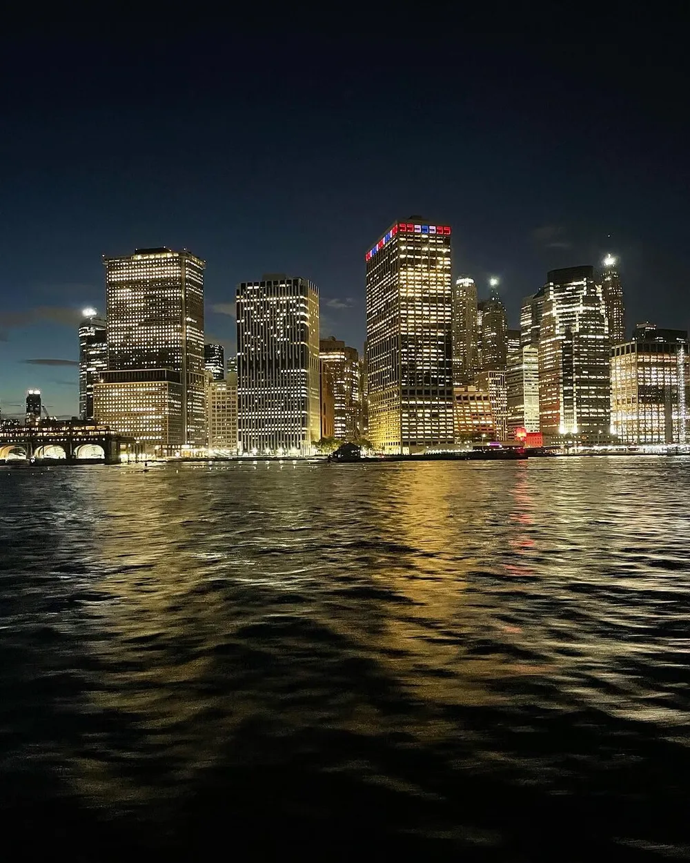 The image captures a glittering skyline of skyscrapers at night reflected in the rippling waters of a large body of water