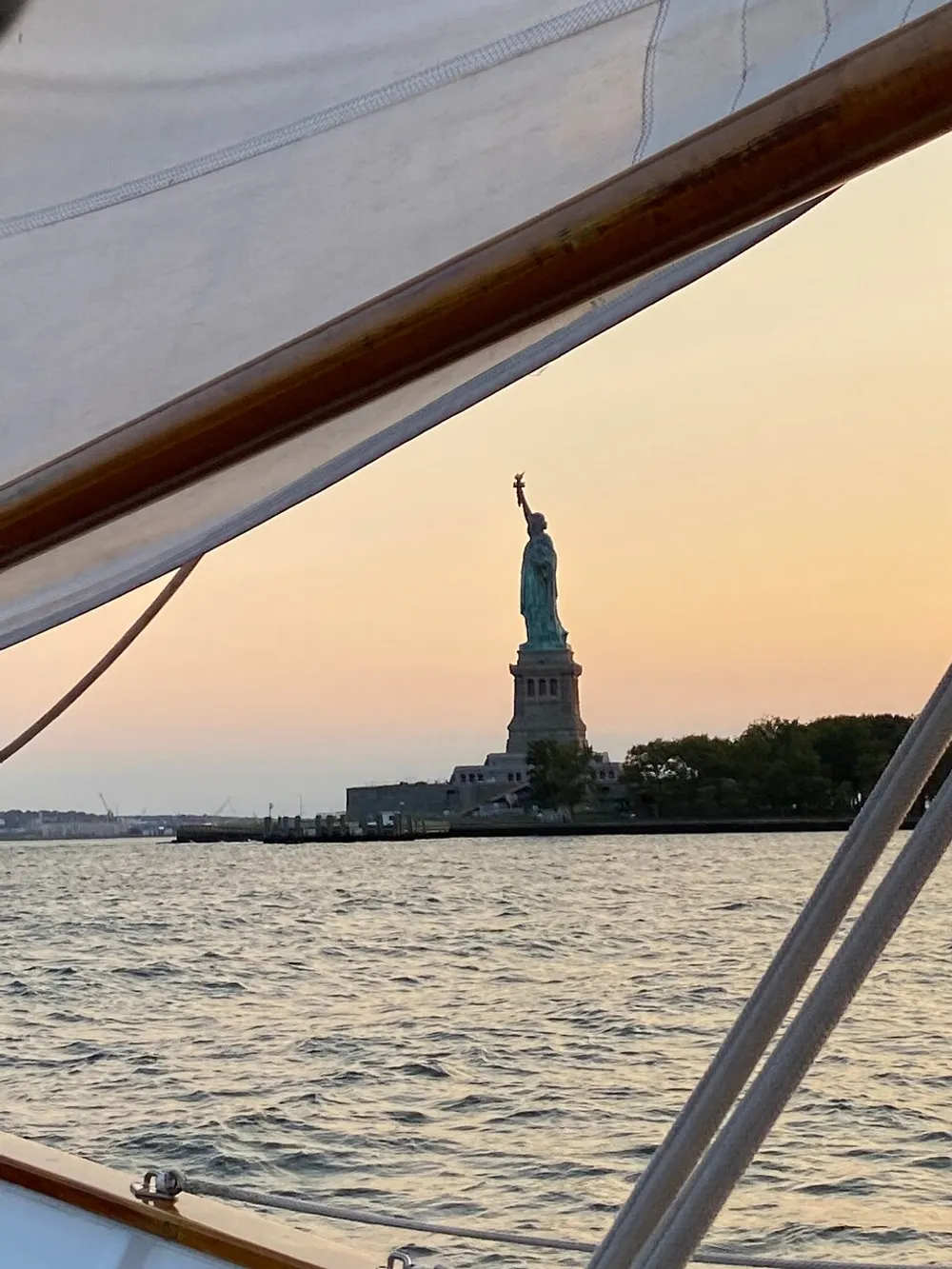 The Statue of Liberty is framed by the sails of a boat during a sunset
