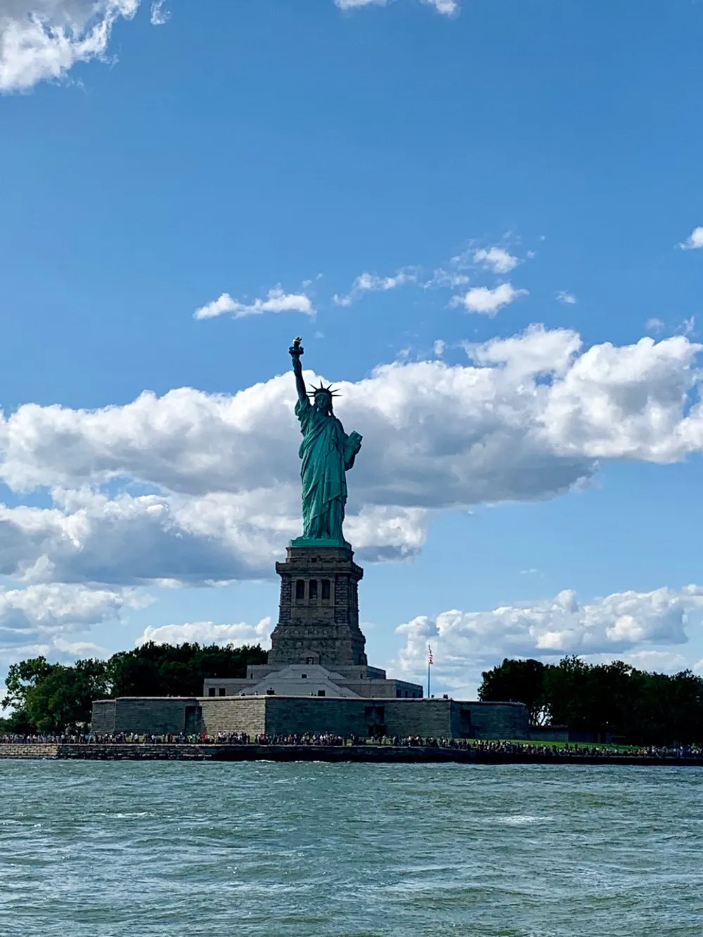 The image shows the Statue of Liberty standing tall against a backdrop of a partly cloudy sky with a crowd of visitors at its base and the waters of New York Harbor in the foreground