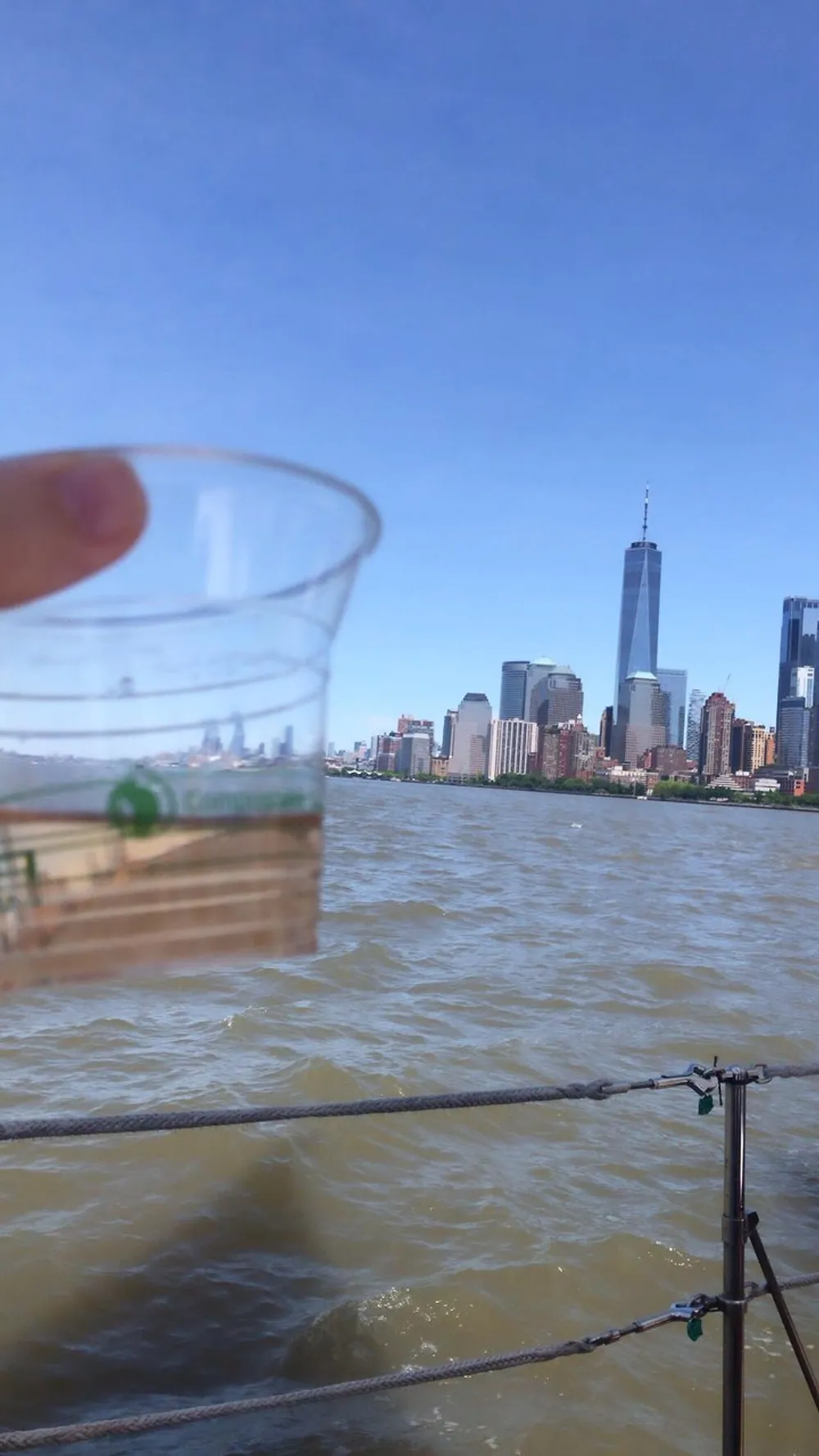 A person holds up a transparent plastic cup partially filled with water against a backdrop of the New York City skyline creating an illusion where the buildings seem to be inside the cup