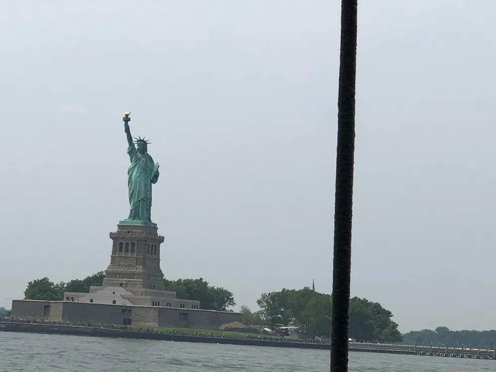The image shows the Statue of Liberty on a cloudy day as viewed from a distance with a rope or cable appearing vertically on the right side