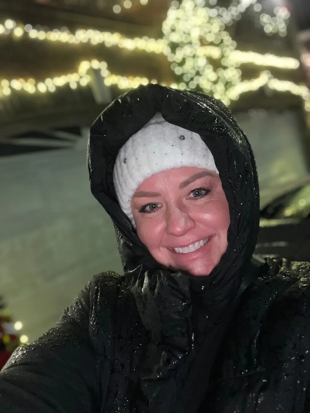 A smiling person in rainwear takes a selfie with twinkling lights in the background on a rainy night