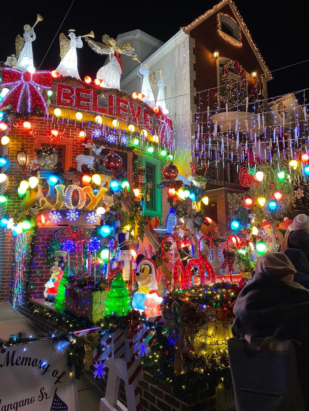 The image shows a house extravagantly decorated with colorful Christmas lights festive ornaments and figures such as angels and Disney characters observed by onlookers in the foreground