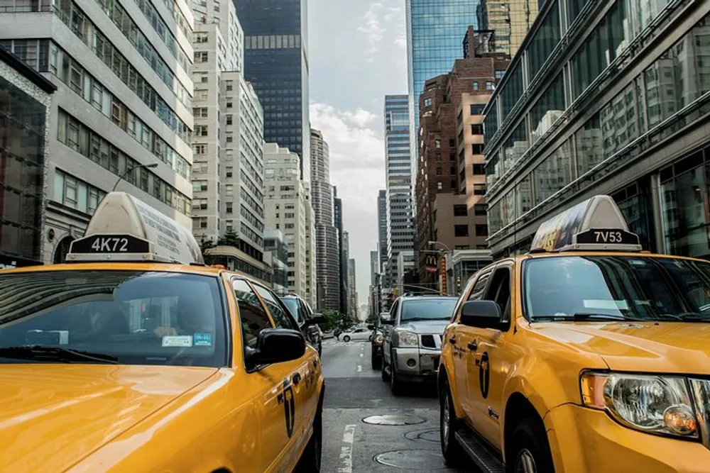 Iconic yellow cabs are lined up in a bustling city street flanked by tall buildings under a cloudy sky