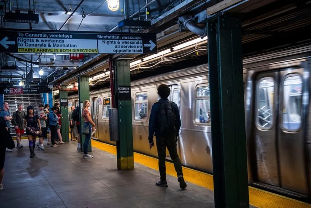 Commuters are waiting on a subway platform as a train arrives in what appears to be a New York City subway station indicated by the signage and setting