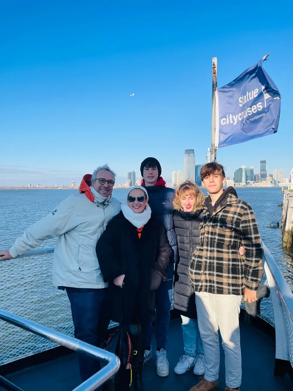 A happy group of people poses for a photo on a boat with the Statue City Cruises flag visible in the background indicating they may be on a tour with a clear blue sky and a coastal city skyline behind them