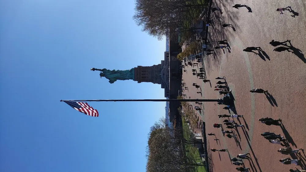 The image is a vertical view of the Statue of Liberty in the distance with an American flag in the foreground and visitors walking around although the photo has been taken with the camera rotated 90 degrees to the left making the scene appear sideways