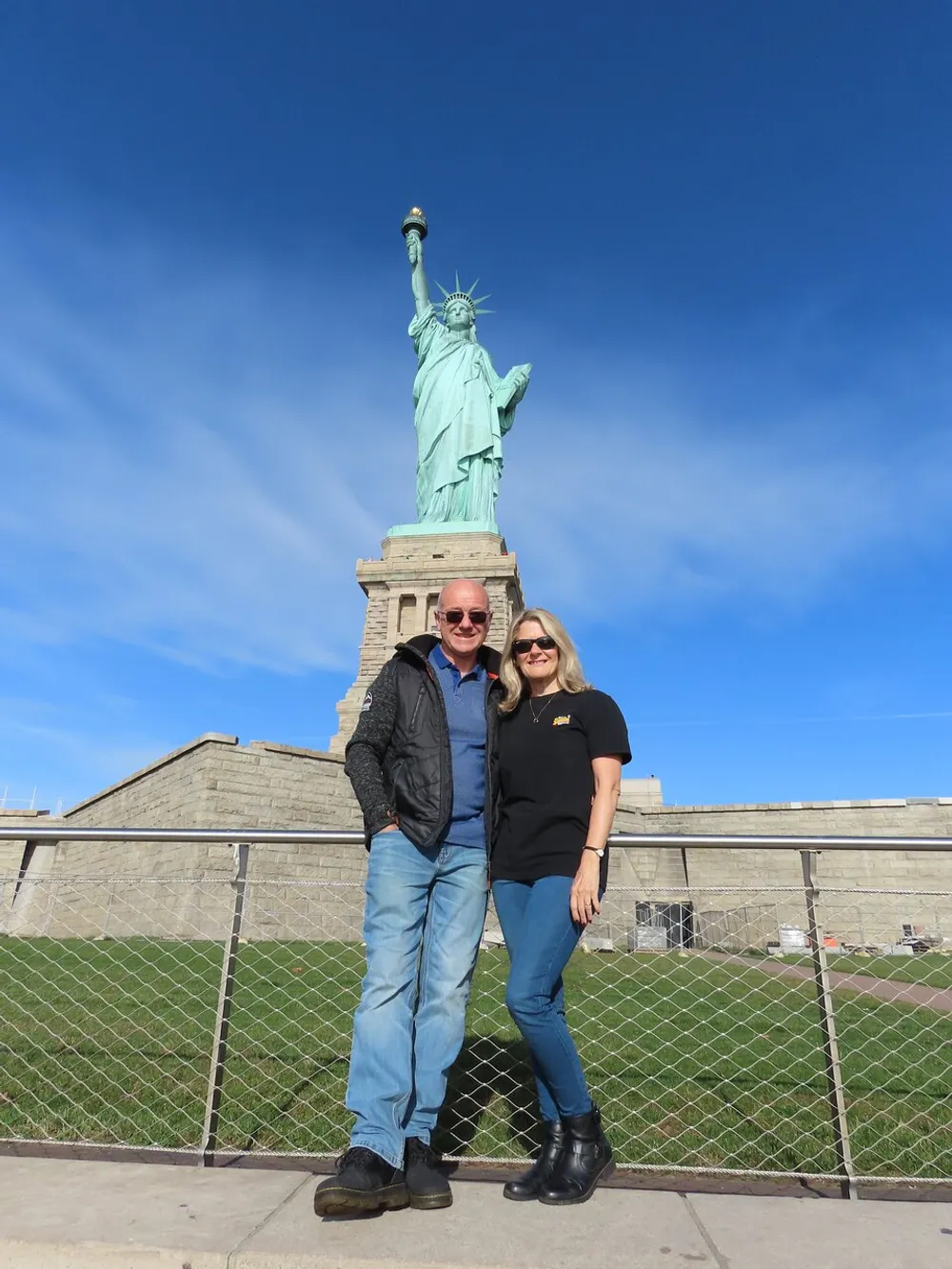 A smiling couple is posing for a photo in front of the Statue of Liberty under a clear blue sky