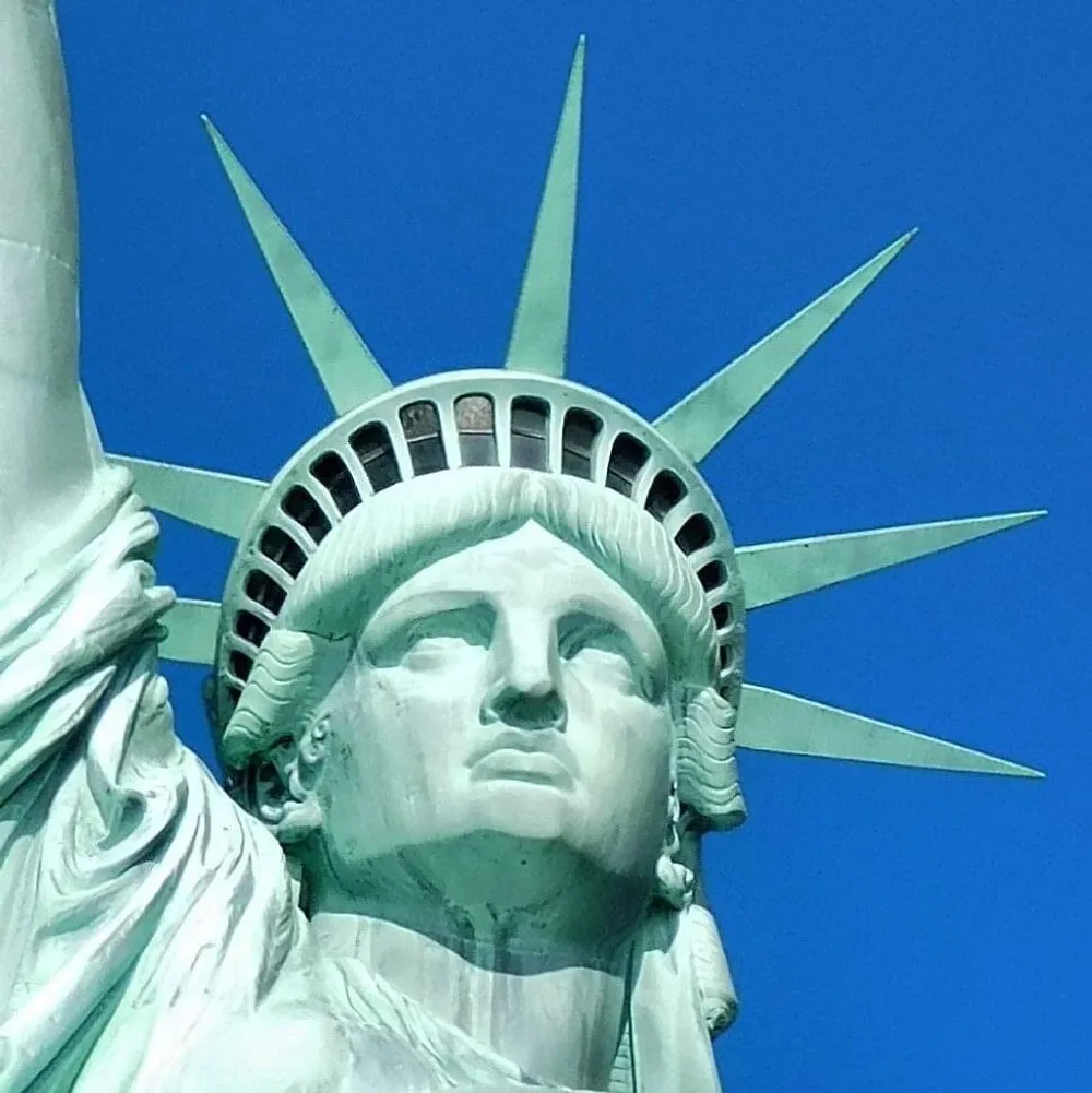 The image shows a close-up of the Statue of Libertys head and crown against a clear blue sky