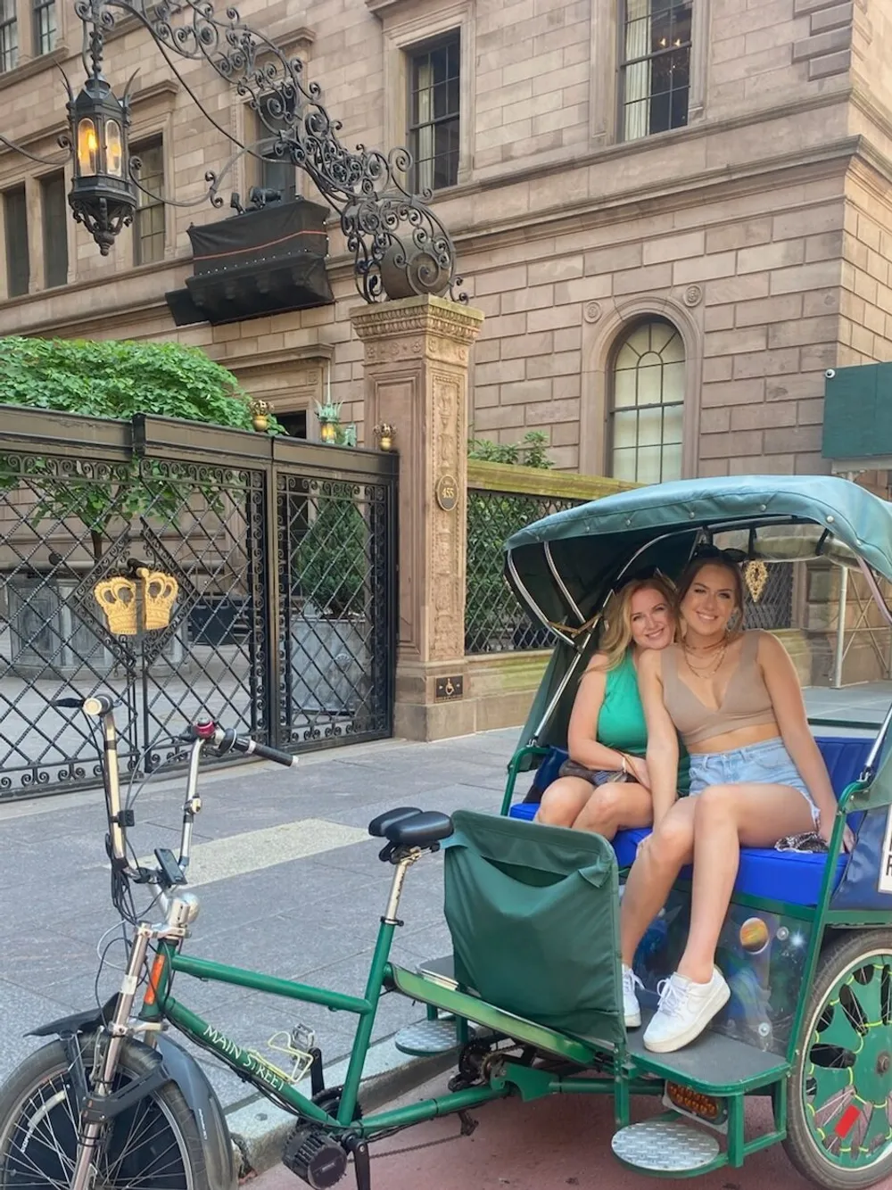 Two people are smiling while seated in a green pedicab parked in front of an elegant building with ironwork detailing