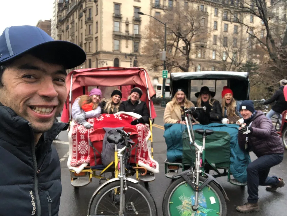 A smiling man takes a selfie in front of two pedicabs carrying cheerful passengers possibly touring a city
