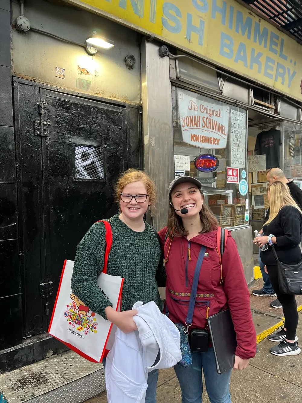 Two smiling young individuals are standing in front of the Yonah Schimmel Knish Bakery possibly after having visited the shop