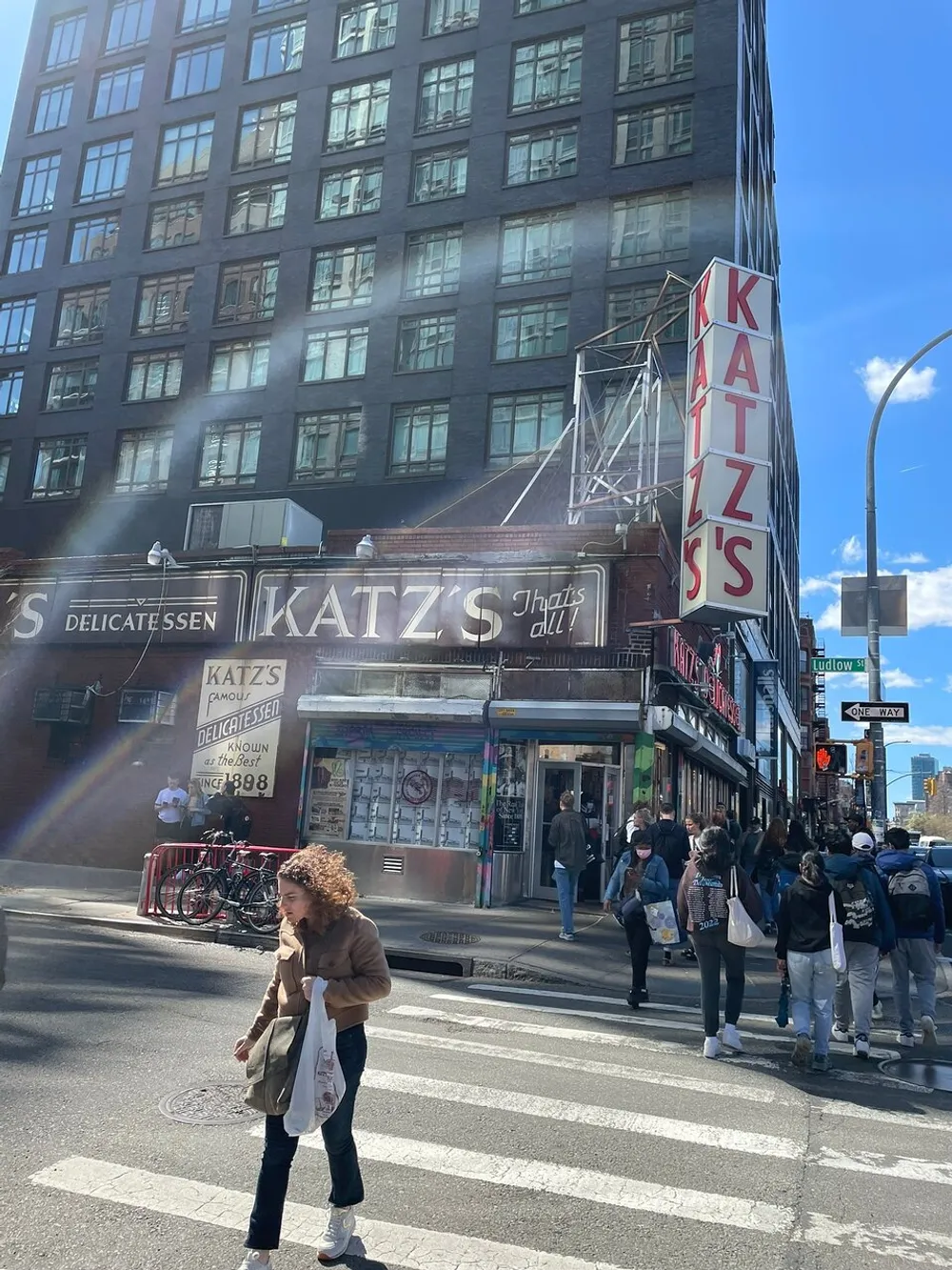 A busy street scene in front of Katzs Delicatessen featuring pedestrians and a prominent red and white sign under clear blue skies