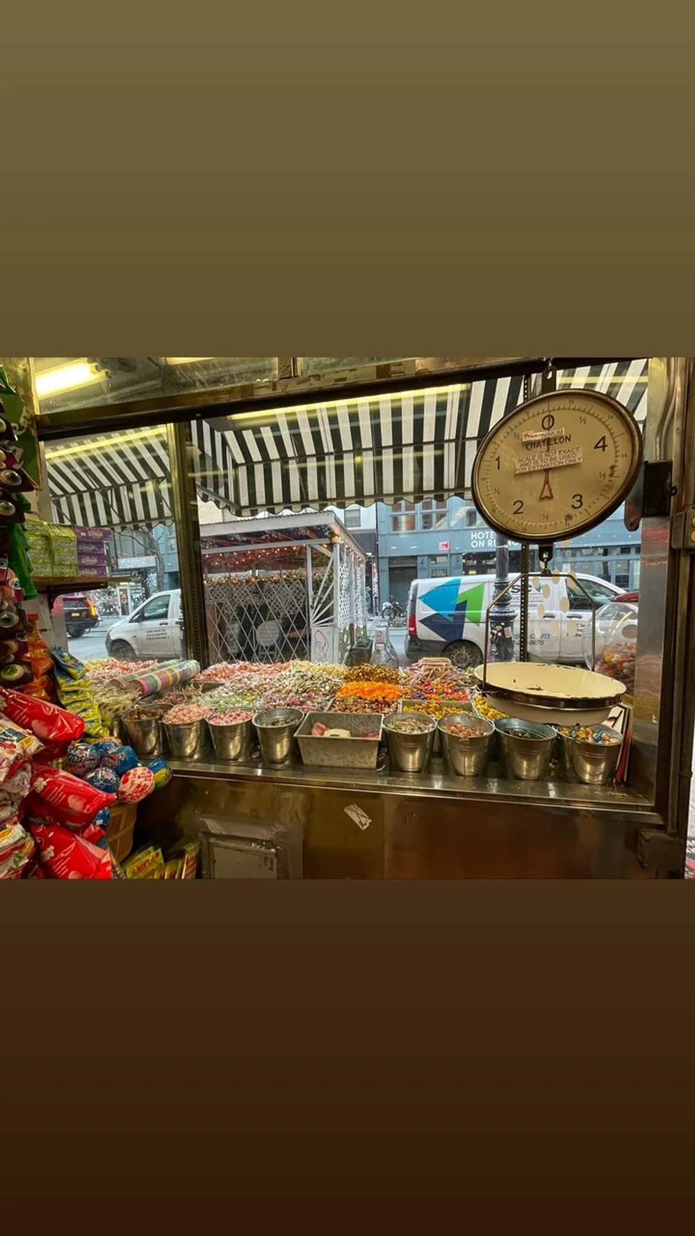 A street vendors stall displays a colorful array of sweets and snacks under the glow of artificial lighting with a bustling city backdrop seen through the service window