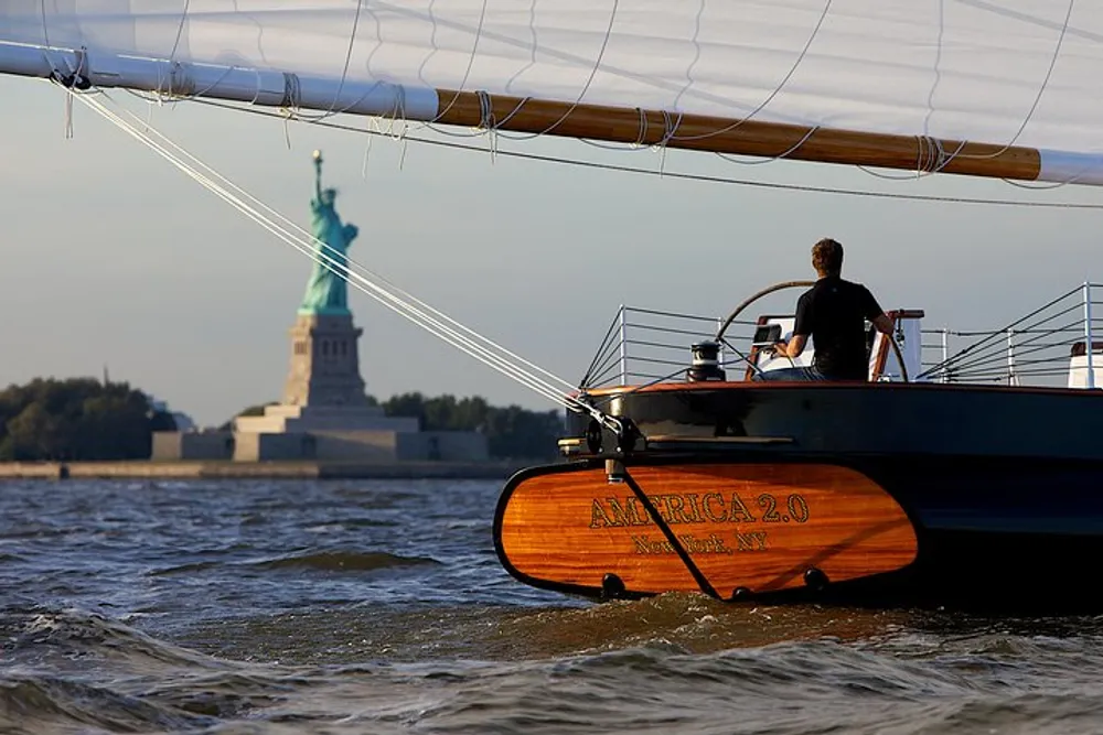 A person is sailing on a boat named America 20 with the Statue of Liberty in the background