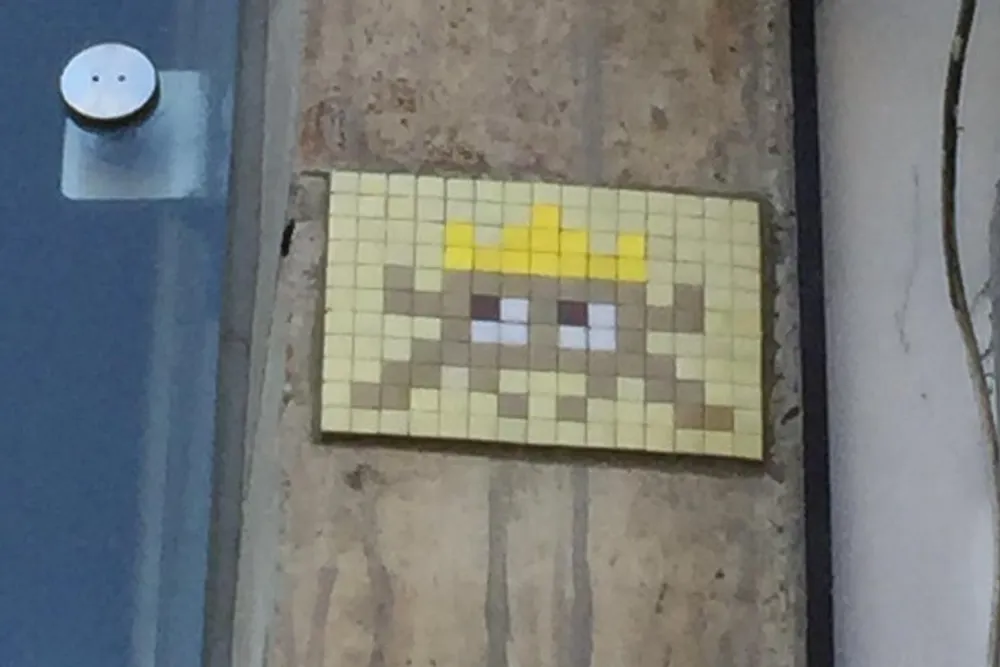 The image shows a pixelated artwork resembling a mosaic depicting a character made of small square tiles mounted on an urban wall