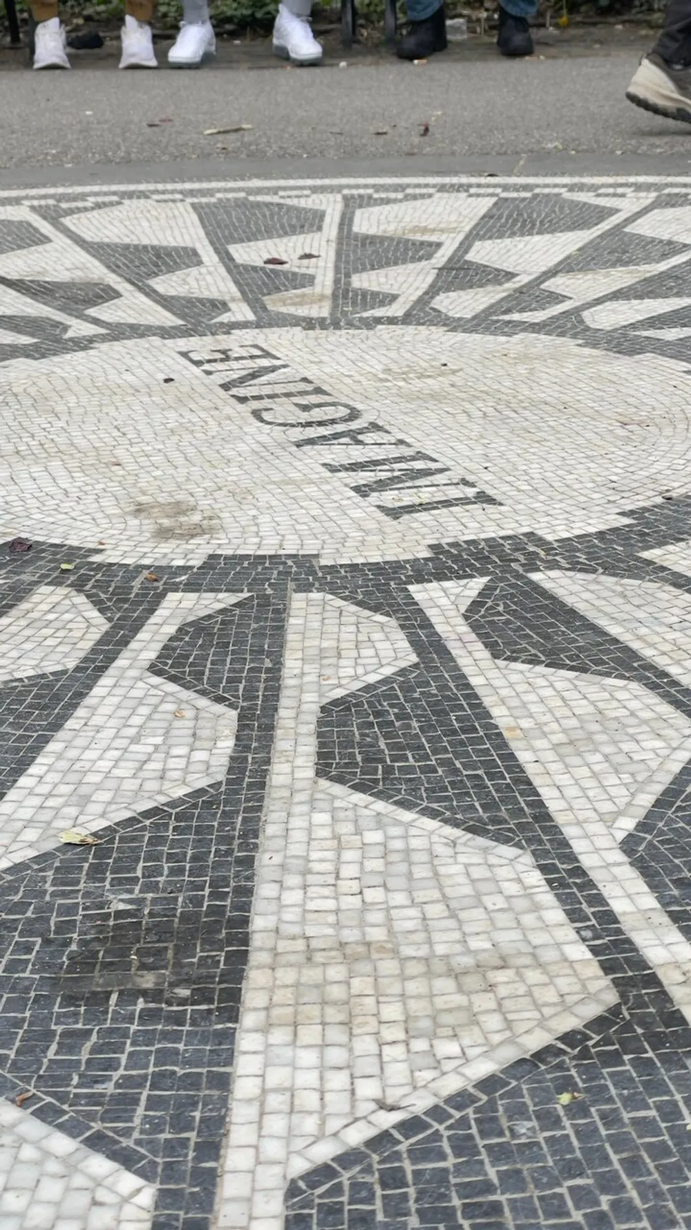 This image shows a detailed mosaic pavement with geometric patterns and the word Imagine prominently displayed with the lower half of the photo revealing the feet of people walking by